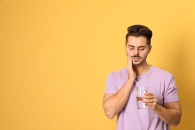 Emotional young man with sensitive teeth and glass of water on color background. Space for text