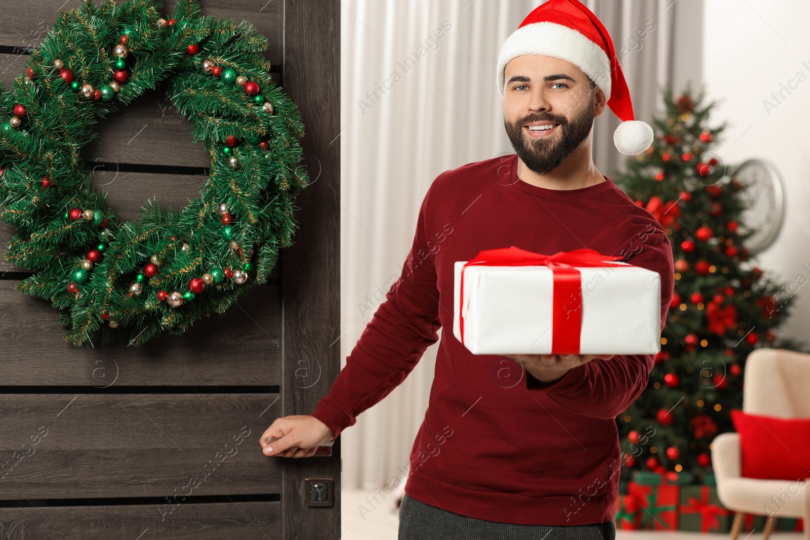 Photo of Young man in Santa hat with Christmas gift box indoors. Sending present by mail