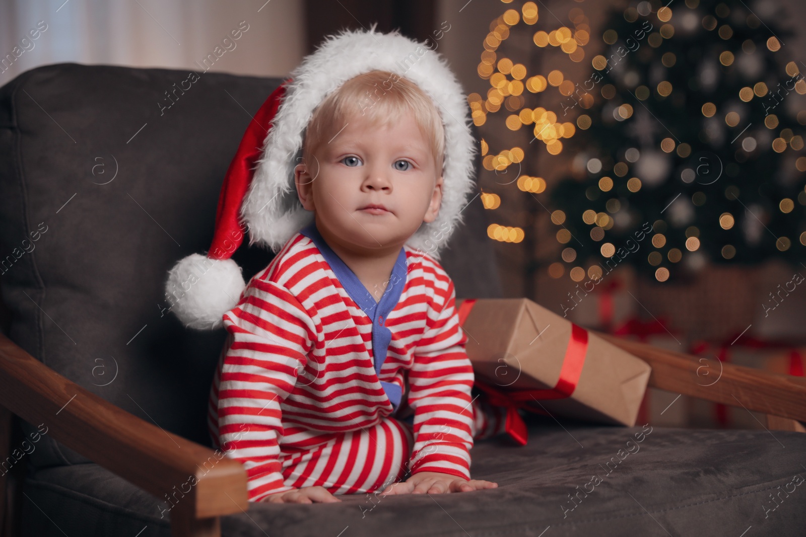 Photo of Baby in Christmas pajamas and Santa hat with gift box sitting in armchair at home