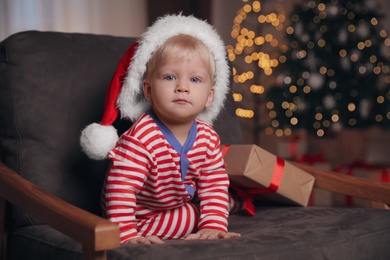 Baby in Christmas pajamas and Santa hat with gift box sitting in armchair at home