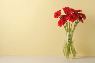 Photo of Bouquet of beautiful red gerbera flowers glass in vase on white table near beige wall. Space for text