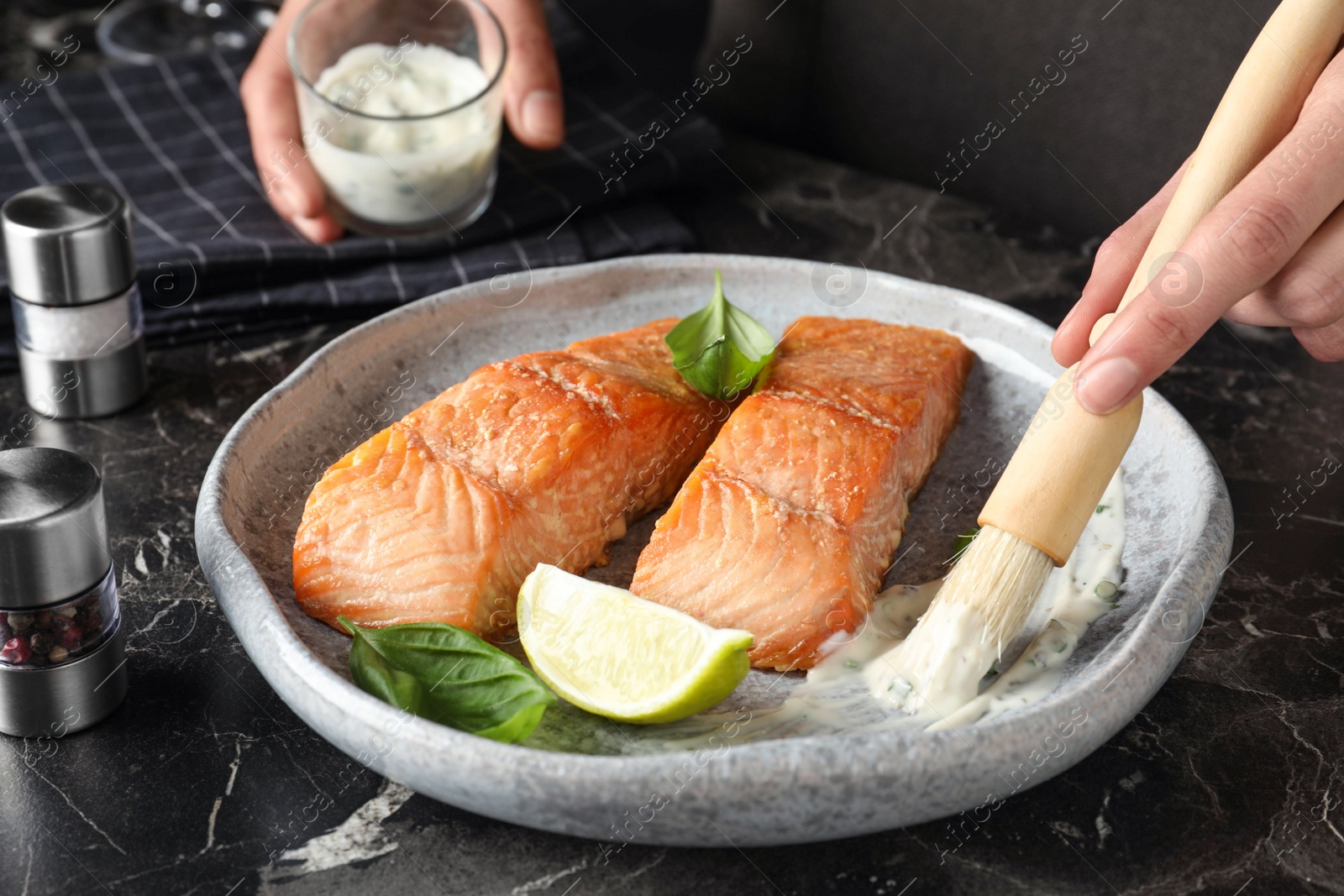 Photo of Woman adding sauce to cooked red fish on dark marble table, closeup