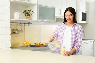 Photo of Beautiful woman pouring lemon water into glass from jug in kitchen. Space for text