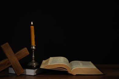 Photo of Church candle, Bible and cross on wooden table against black background, space for text