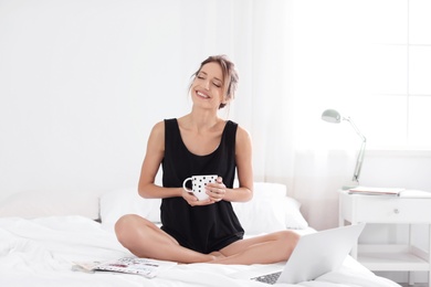 Photo of Young happy woman with cup of aromatic coffee sitting on bed at home. Lazy morning