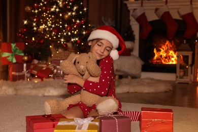 Photo of Cute child in Santa hat hugging toy bunny near Christmas gifts on floor at home
