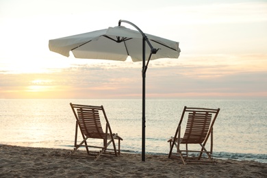 Photo of Wooden deck chairs and outdoor umbrella on sandy beach at sunset. Summer vacation
