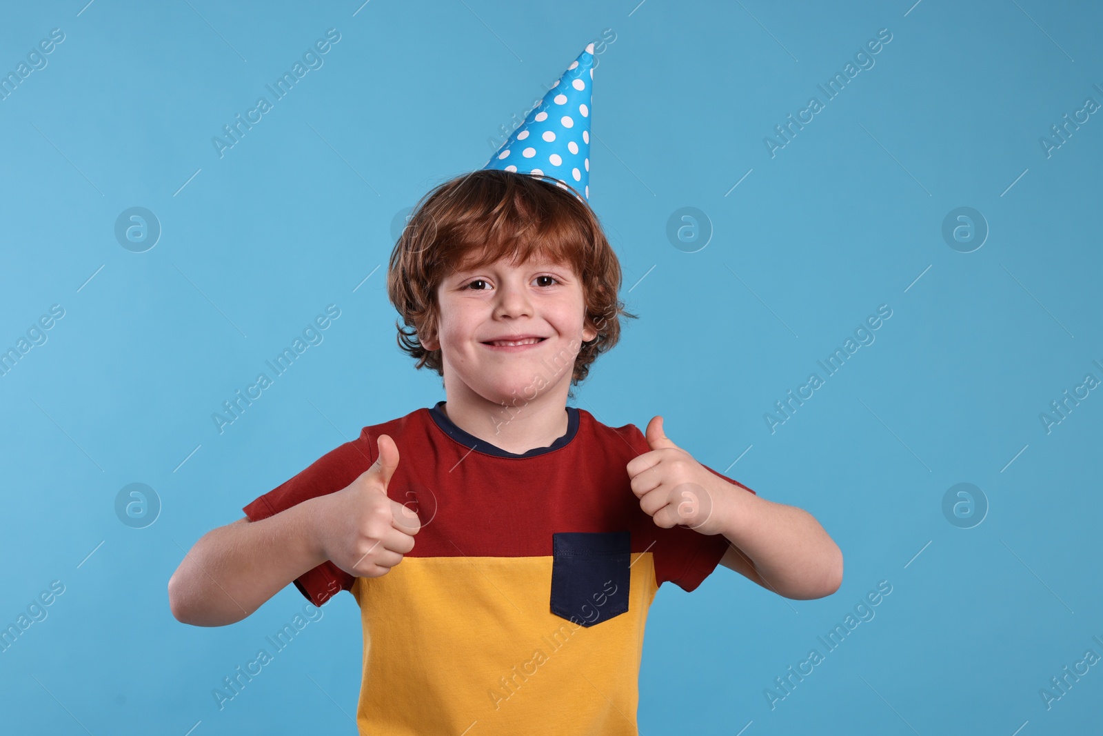 Photo of Birthday celebration. Cute little boy in party hat showing thumbs up on light blue background