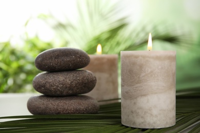 Photo of Burning candles and spa stones with palm leaf on table against blurred green background