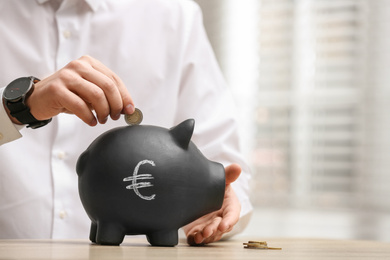 Man putting coin into piggy bank with euro sign at wooden table, closeup. Space for text