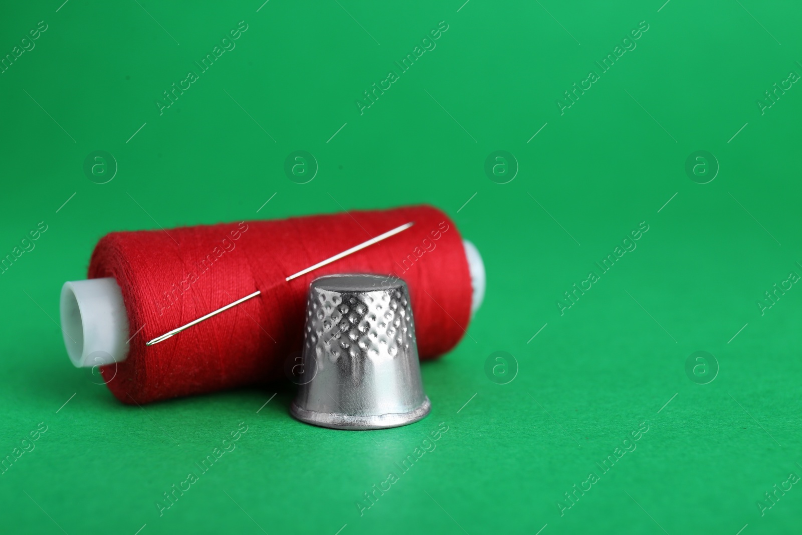 Photo of Silver thimble, thread and needle on green background, space for text. Sewing accessories