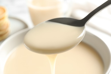 Photo of Spoon of pouring condensed milk over bowl, closeup. Dairy products