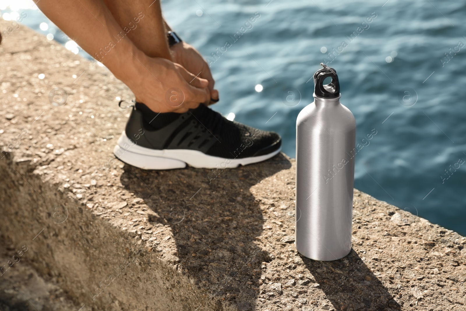 Photo of Young man tying shoelaces near bottle of water at riverside on sunny day