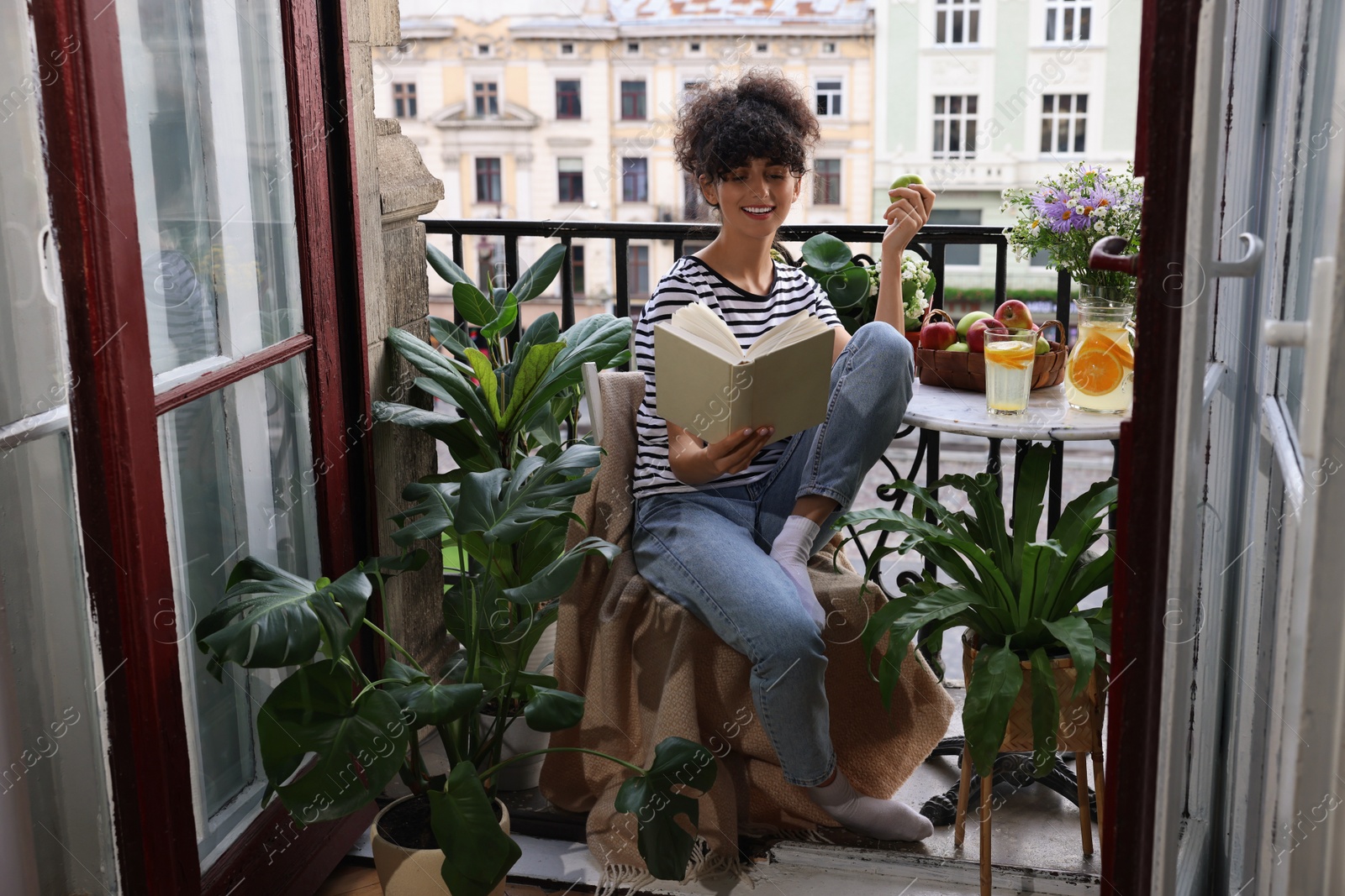 Photo of Young woman reading book at table on balcony with beautiful houseplants