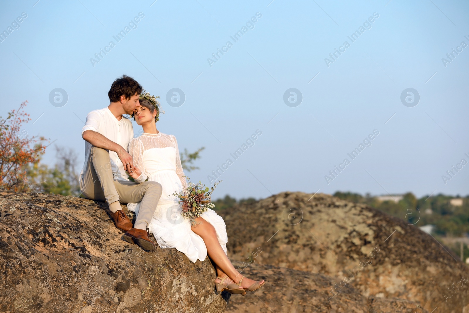 Photo of Happy newlyweds with beautiful field bouquet sitting on rock outdoors