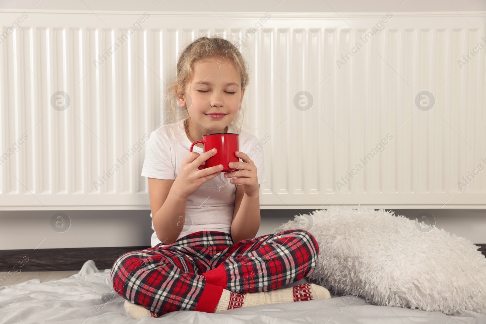 Photo of Little girl with cup of hot drink near heating radiator indoors