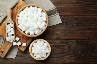 Photo of White sugar cubes in bowls and scoop on wooden table, flat lay. Space for text