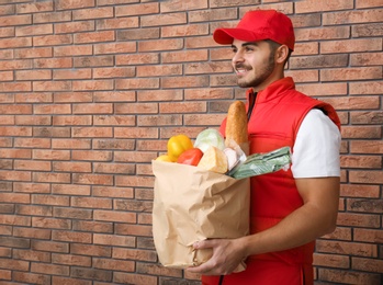 Photo of Food delivery courier holding paper bag with products near brick wall. Space for text