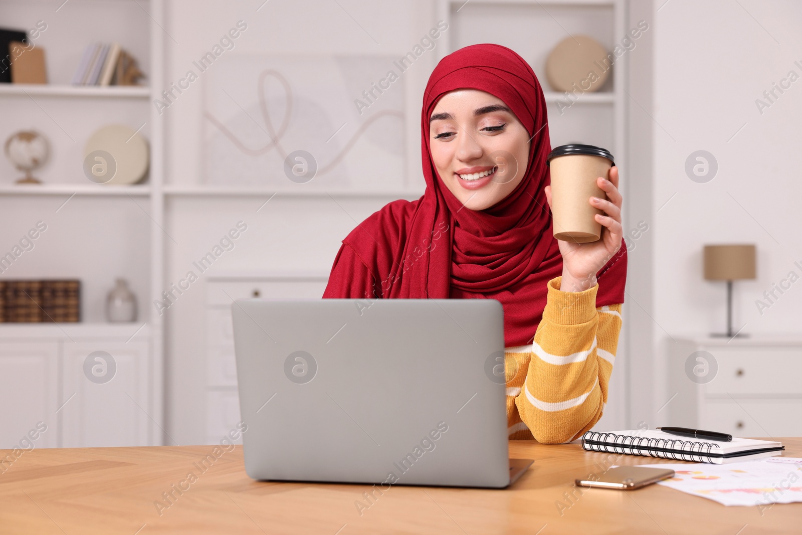 Photo of Muslim woman in hijab with cup of coffee using laptop at wooden table in room