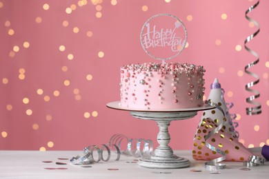 Photo of Beautiful birthday cake and decor on white table against blurred festive lights, space for text