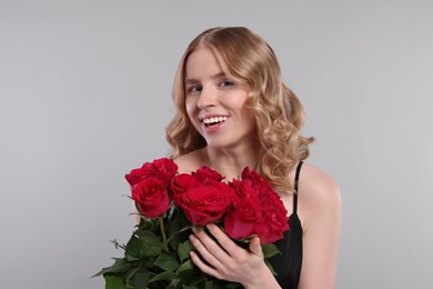 Beautiful woman with blonde hair holding bouquet of red roses on light grey background