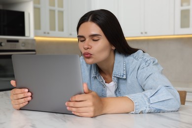 Photo of Young woman having video chat via laptop and blowing kiss at table in kitchen