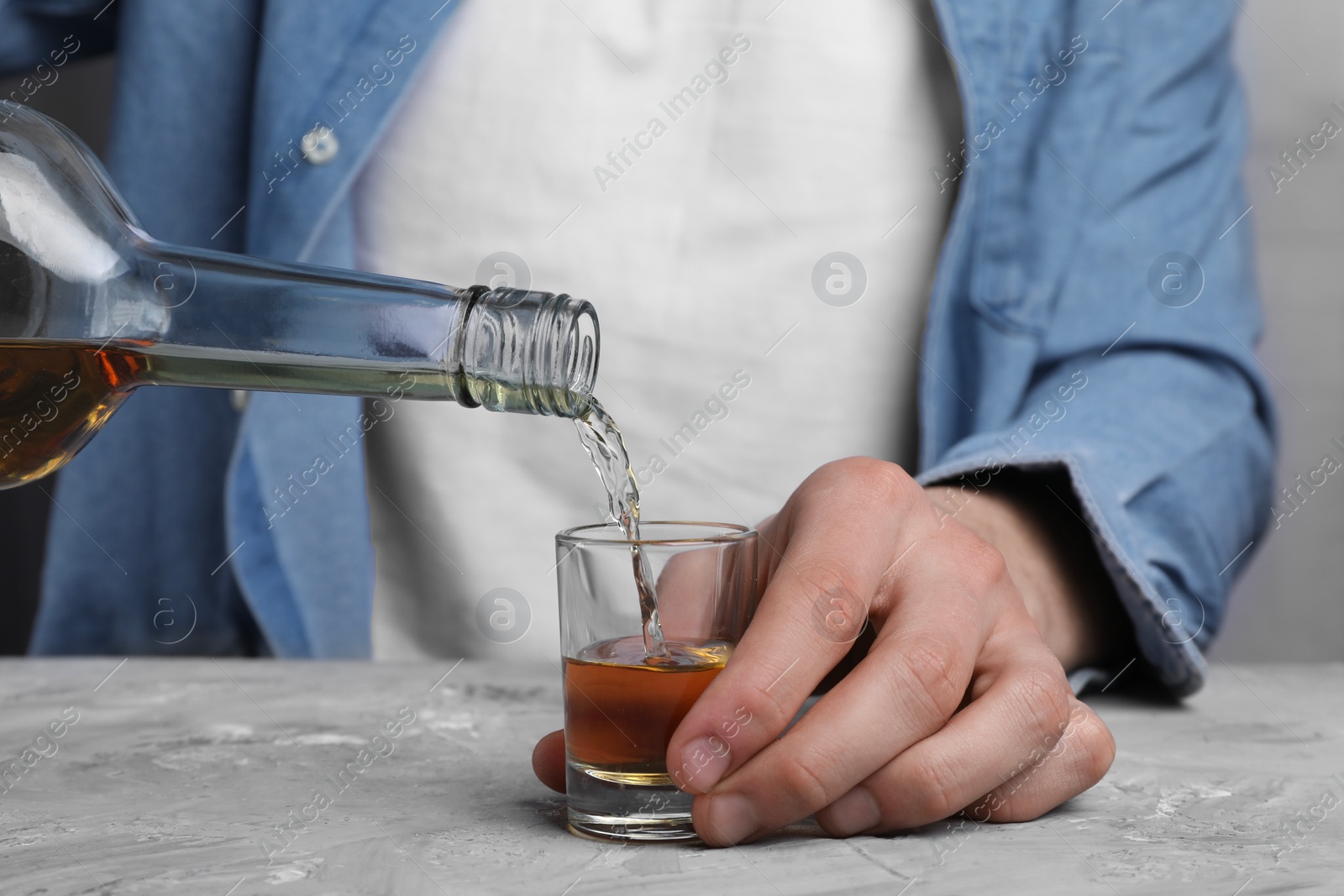 Photo of Alcohol addiction. Man pouring whiskey from bottle into glass at grey textured table, closeup