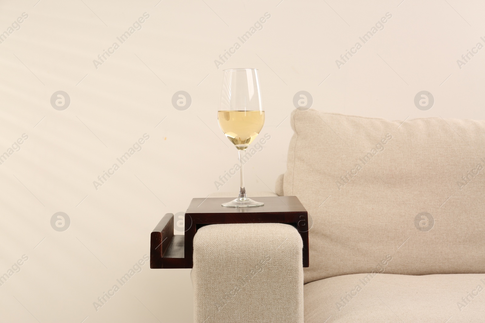 Photo of Glass of white wine on sofa with wooden armrest table indoors. Interior element