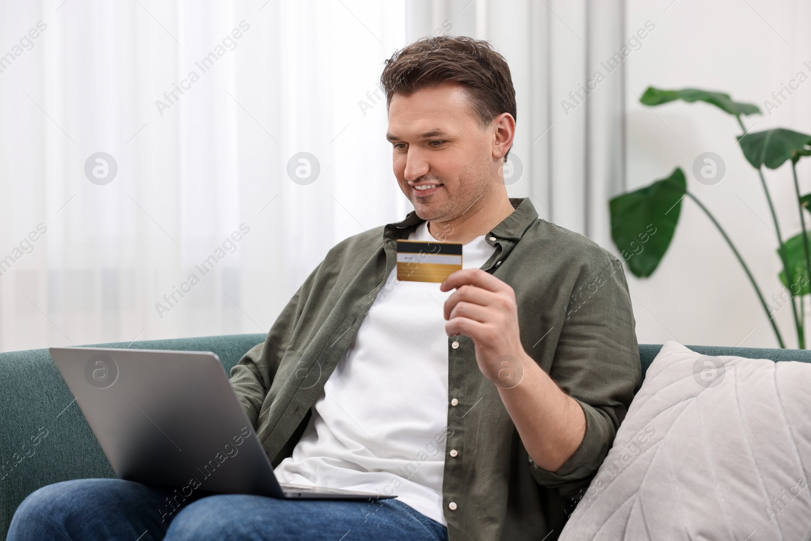 Photo of Happy man with credit card using laptop for shopping online on sofa at home