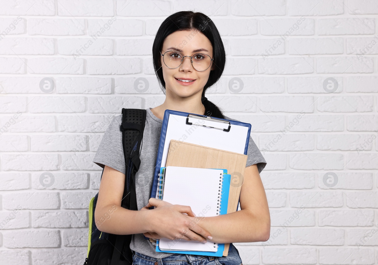 Photo of Smiling student with notebooks, clipboard and backpack near white brick wall
