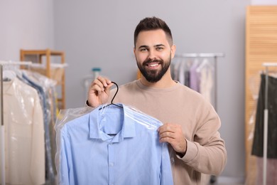 Dry-cleaning service. Happy man holding hanger with shirt in plastic bag indoors