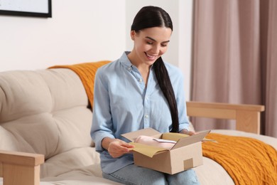 Happy woman opening parcel with greeting card on sofa at home. Christmas gift