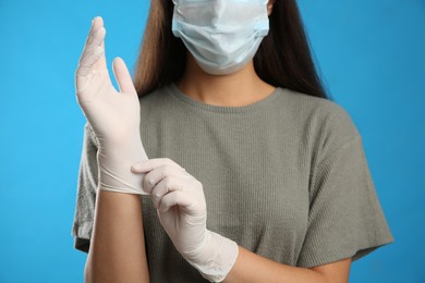 Photo of Woman in protective face mask putting on medical gloves against blue background, closeup
