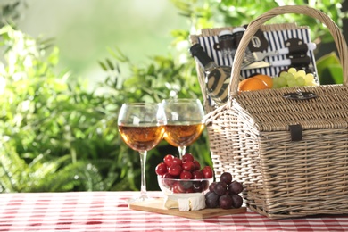 Wicker picnic basket with different products on checkered tablecloth against blurred background, space for text