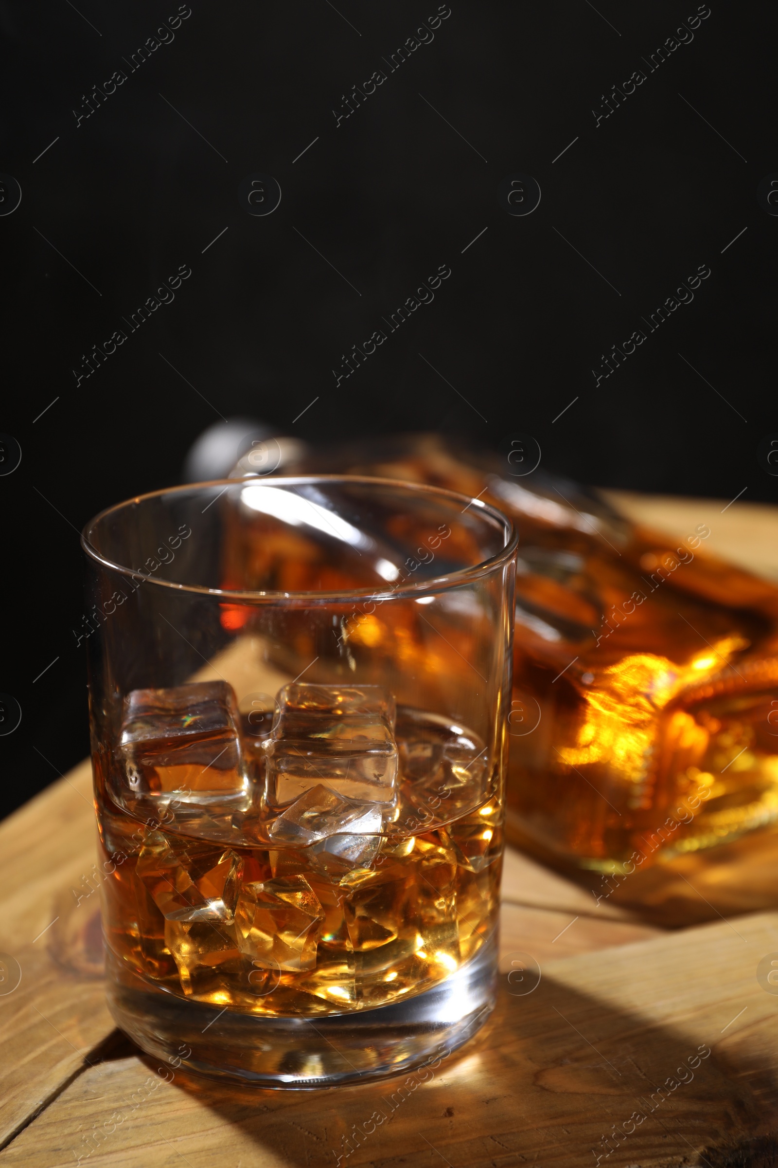 Photo of Whiskey with ice cubes in glass and bottle on wooden crate against black background, closeup