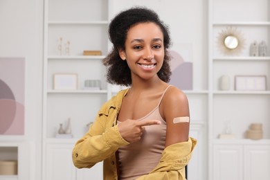 Happy young woman pointing at adhesive bandage after vaccination indoors