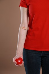 Photo of Blood donation concept. Woman with adhesive plaster on arm holding red heart against brown background, closeup