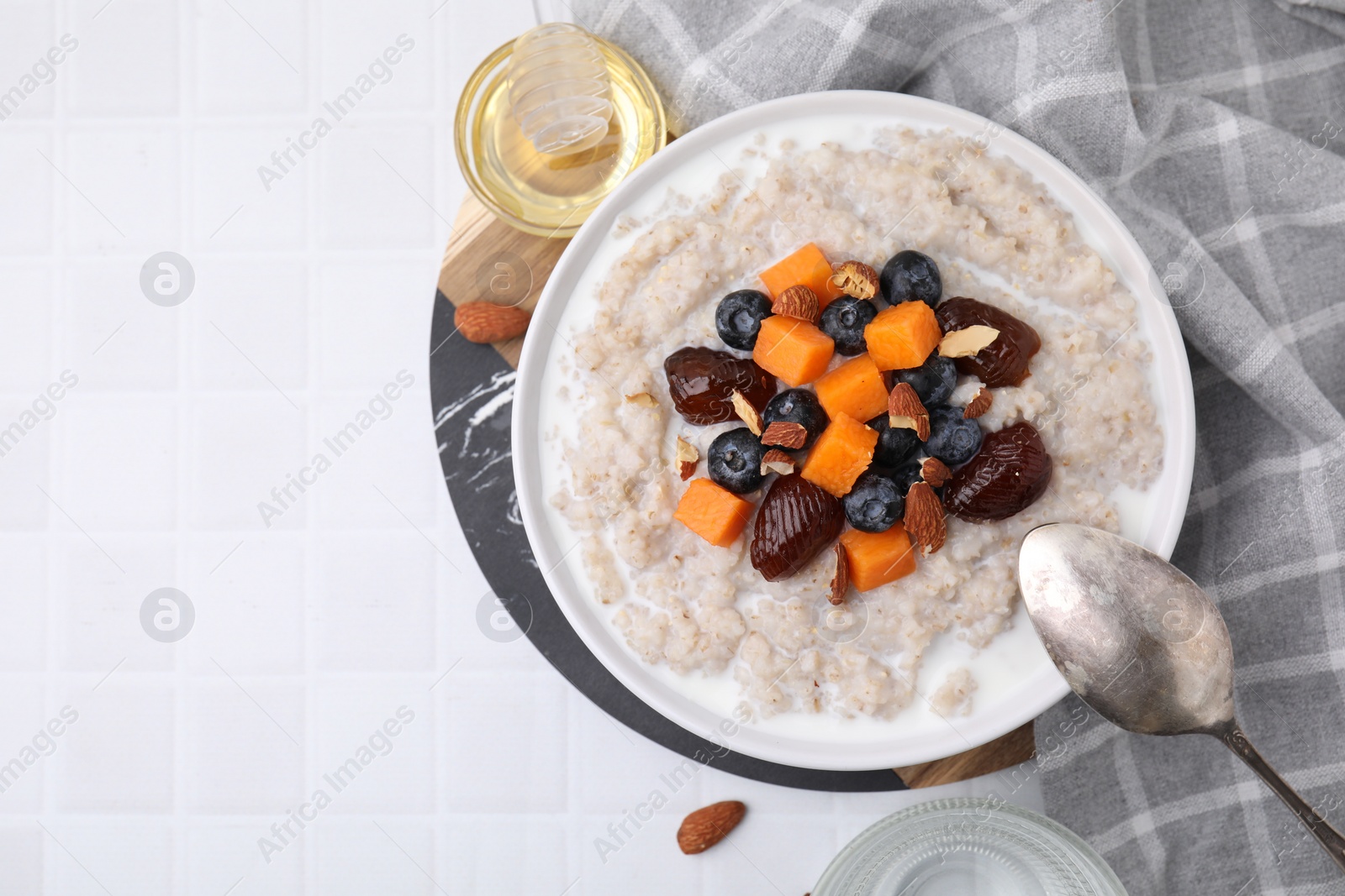 Photo of Delicious barley porridge with blueberries, pumpkin, dates and almonds served with honey on white tiled table, flat lay. Space for text