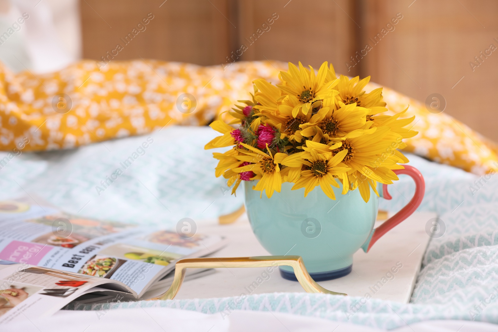 Photo of Beautiful bright flowers in cup, open magazine and tray on bed. Space for text