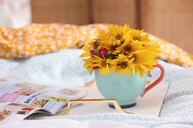 Photo of Beautiful bright flowers in cup, open magazine and tray on bed. Space for text