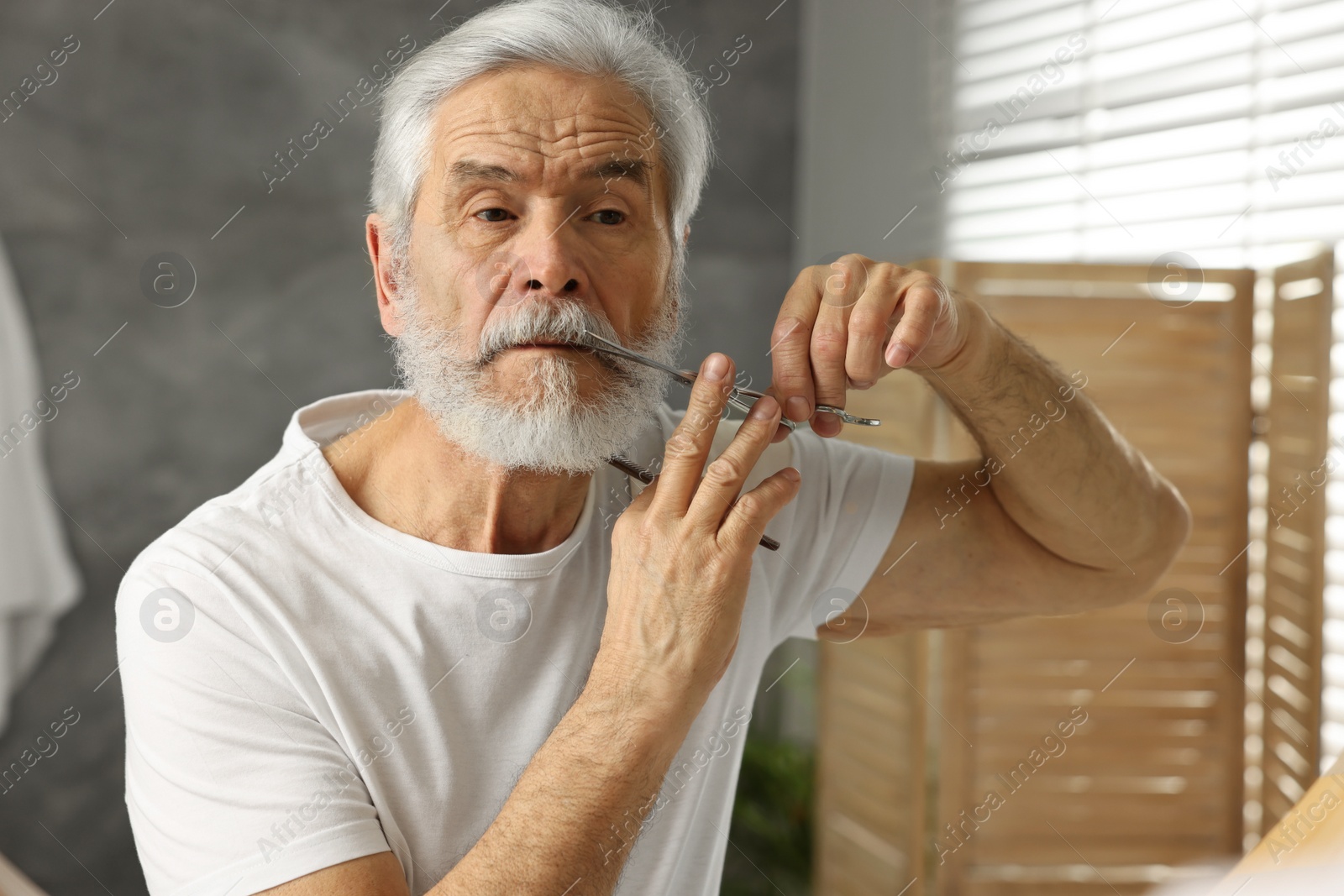 Photo of Senior man trimming mustache with scissors near mirror in bathroom