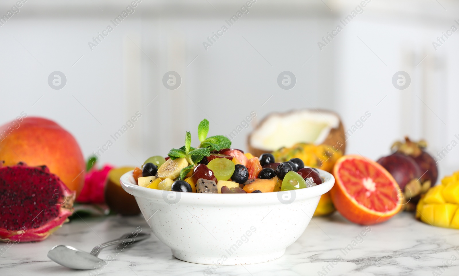 Photo of Delicious exotic fruit salad and ingredients on white marble table in kitchen