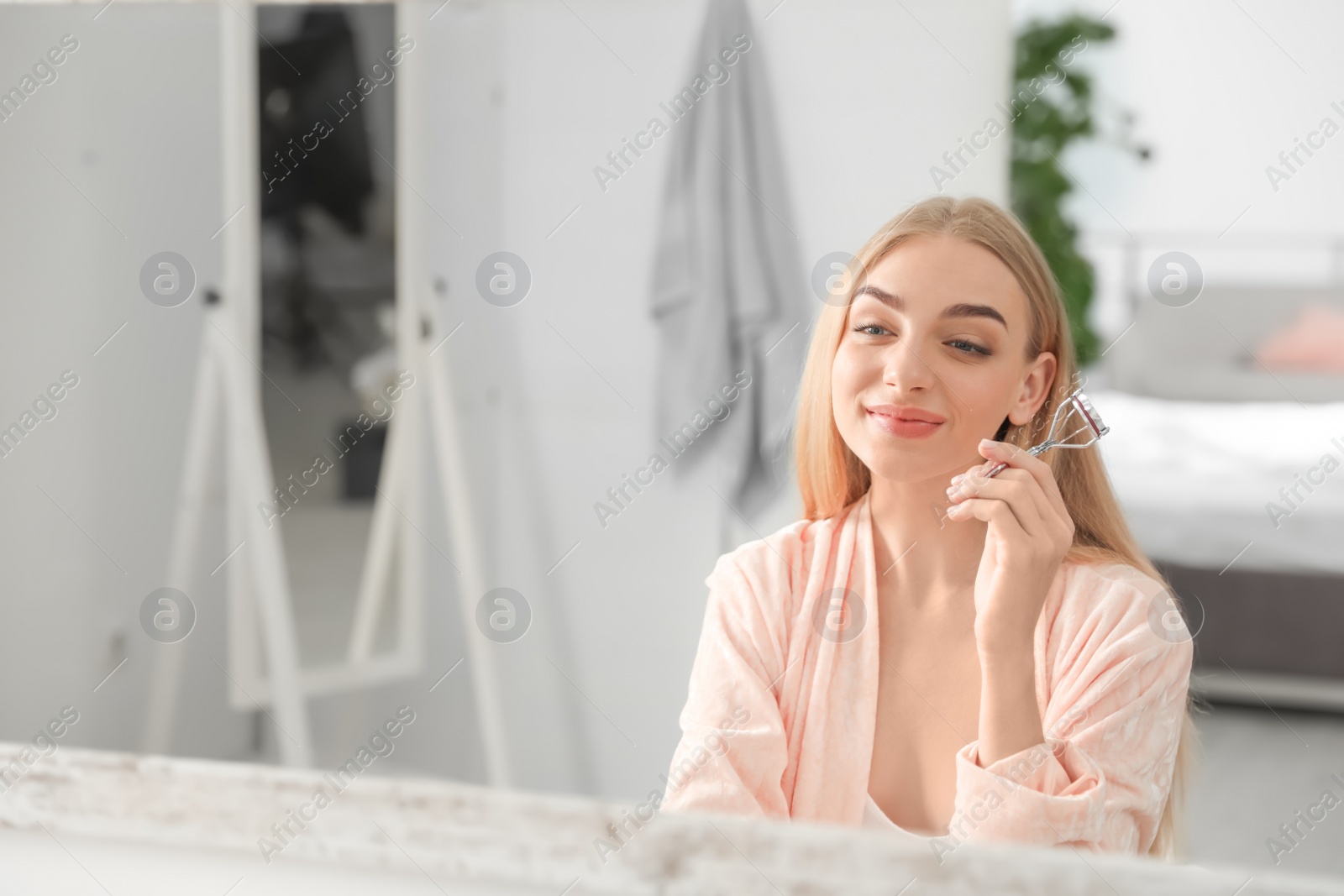 Photo of Young woman curling her eyelashes near mirror indoors