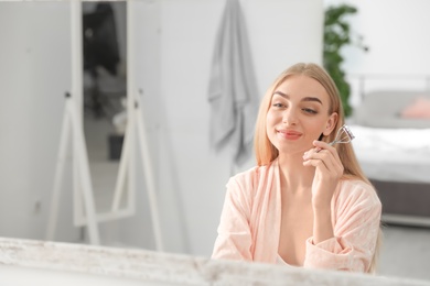 Young woman curling her eyelashes near mirror indoors