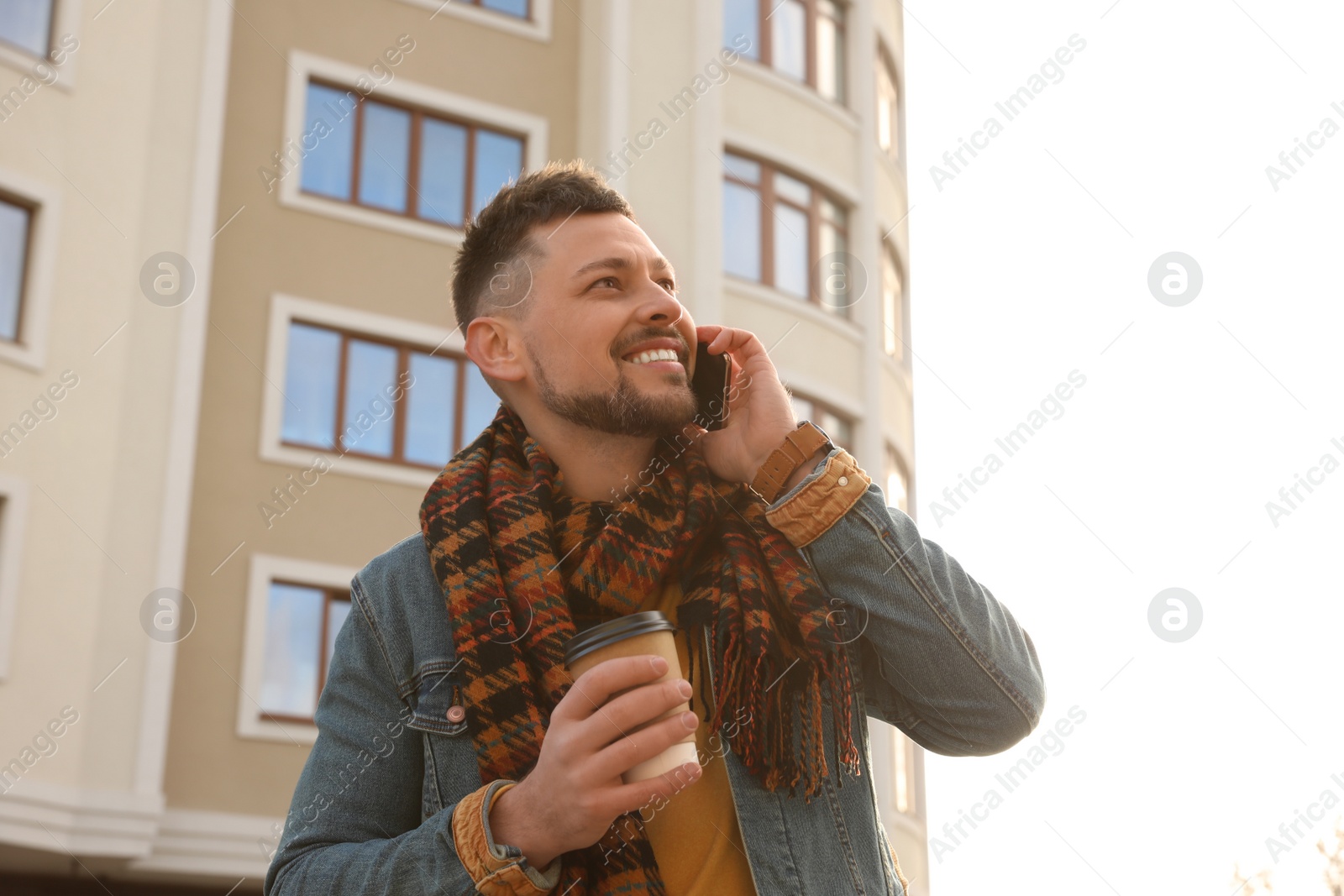 Photo of Man with cup of coffee talking on smartphone on city street in morning