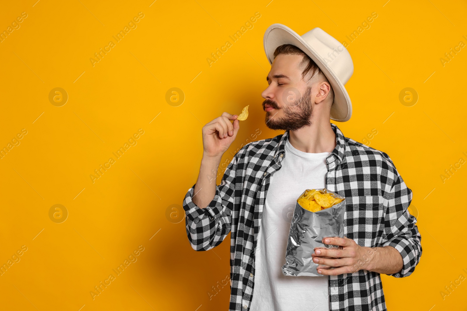 Photo of Handsome young man with bag of tasty potato chips on yellow background. Space for text
