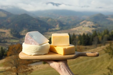 Photo of Woman holding board with different types of delicious cheeses against mountain landscape, closeup
