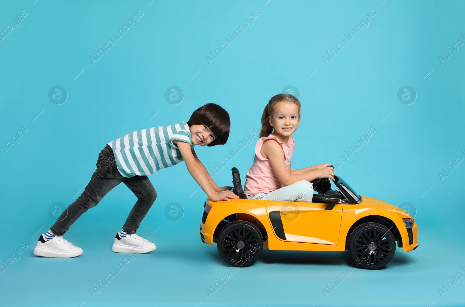 Photo of Cute boy pushing children's electric toy car with little girl on light blue background