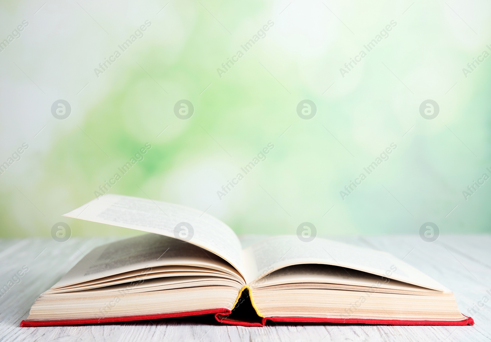 Photo of Open book on white wooden table against blurred green background. Space for text