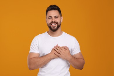 Photo of Thank you gesture. Happy grateful man holding hands near heart on orange background